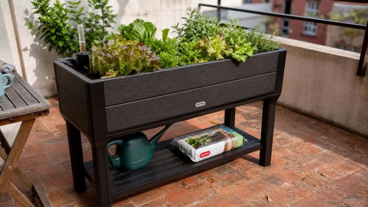 Mesa de cultivo elevada de resina oscura plantada con lechugas y aromáticas en una terraza soleada, con una regadera y un saco de sustrato guardados en el estante inferior